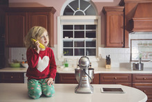 Cute Girl Playing With Banana At Table By Appliance In Kitchen