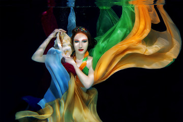 Red hair girl with big sea shell in colorful clothes on the dark background underwater