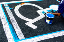 Close Up Of Man Painting Empty Handicapped Reserved Parking Space With Wheelchair Symbol On Black Asphalt. No Parking White Painted Letters And Blue Diagonal Lines In Background.