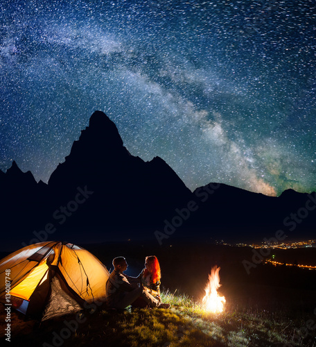 Night Camping Romantic Couple Sitting Face To Face In Front Tent Near Campfire Under Shines Starry Sky On The Background Silhouettes Of The High Mountains And Luminous Village In The Valley
