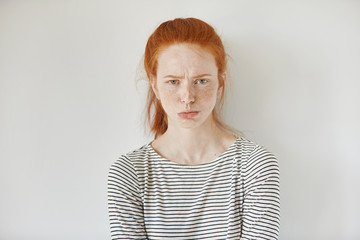 Pretty girl with ginger hair, gathered in ponytail pouting her lips, looking at camera with irritated and offended face expression while standing isolated against white studio wall background