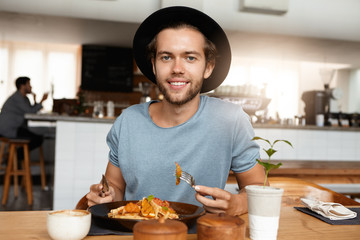 People, lifestyle, health and nutrition. Handsome young bearded male eating healthy food with knife and fork during lunch break, sitting at wooden cafe table with plate and mug and enjoying vegetables