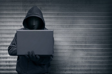 Wall Mural - Hooded hacker with mask holding laptop while typing