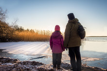 Woman And Young Girl Looking At Beautiful Winter Sunset Against Icy Water And Blue Sky, Late Afternoon.