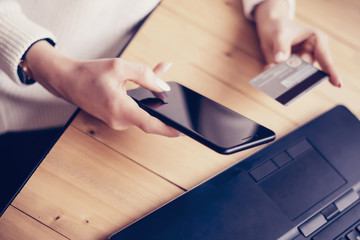 Closeup of young woman making online shopping by laptop and smartphone at working place.Girl touching home button on mobile phone, writing security number credit card.Horizontal, blurred background.