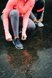 Detail of urban athletes lacing sport footwear for running over asphalt under the rain. Two women getting ready for outdoor training and fitness exercising on cold winter weather.
