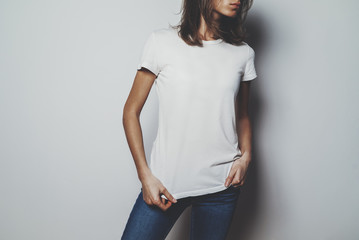 Wall Mural - Young hipster girl wearing blue jeans and white blank t-shirt with space for your logo or design, mock-up of white cotton t-shirt, white wall in the background