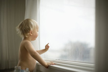 Shirtless Young Toddler Looking Out The Window Of His Home.
