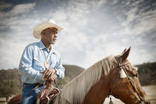 Man Wearing A Cowboy Hat Sits On A Horse Resting His Hands On The Pommel And Looks Away With A Watchful Gaze.