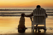 © Erickson Stock - Rear view of man with dog sitting in deck chair on beach during sunset
