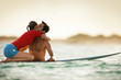 © Erickson Stock - Woman kneeling on a surf-board, hugging her boyfriend.