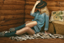 Portrait Of Gorgeous Long-legged Girl Wearing Jeans Shorts, Shirt, Suede Boots With Floral Embroidery And Stetson Felt Hat Sitting In The Wooden Interior On Plaid. Country Style Concept. Copy-space