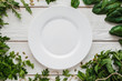 Empty white plate in seasoning frame void flat lay. Top view on blank porcelain dish bordered with fresh green, free space. Cooking, kitchen, menu, spices, cuisine concept