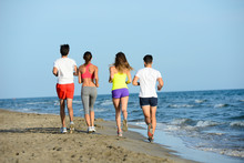 Group Of Young People Running In The Sand On The Shore Of A Beach By The Sea At Sunset During A Sunny Summer Holiday Vacation
