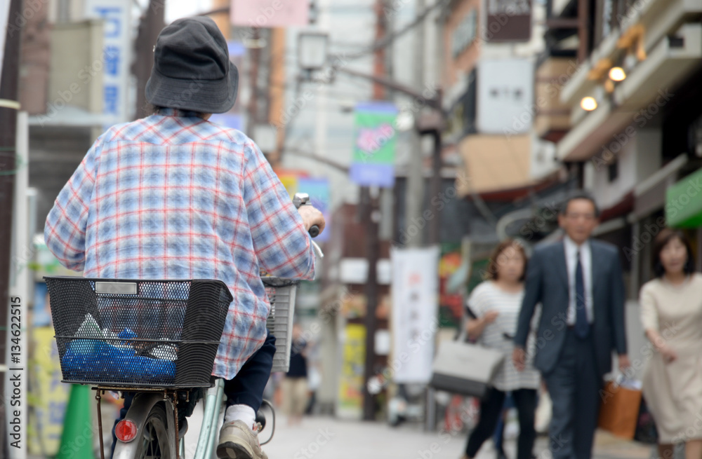 Starsza osoba na rowerze z koszykiem jedzie przez miasto - obrazy, fototapety, plakaty 