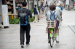 Idący pieszo młody mężczyzna z plecakiem oraz kobieta z plecakiem jadąca na rowerze z zamontowanym koszykiem do przewożenia dziecka