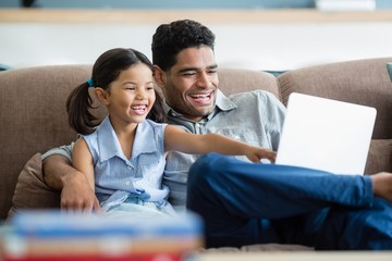 Canvas Print - Father and daughter sitting on sofa and using laptop