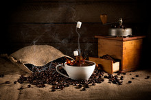 Coffee Art Composition: Scattered Roasted Beans From A Bag, Coffee Grinder With Ground Coffee And A Cup Of Coffee With A Splash Of Falling Sugar. In Fragrant Smoke From Coffee. On Sackcloth