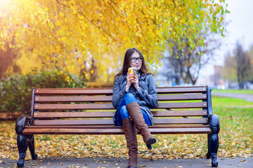 Beautiful young woman sitting on a bench drinking coffee or hot tea in the spring  autumn coat enjoying in park outdoors, glasses, urban life, the concept of breakfast in nature