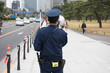 In the picture we can see a Japanese cop announcing and some people are walking in the street and at the background some cars and high rise building can be seen.
