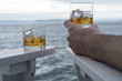Two glasses of whiskey on the rocks by the sea, man holding up dram