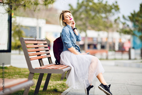 Young Cute Blond Girl With Short Hair Sitting On Wooden Bench And