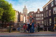 Two Lovers Person In Amsterdam On A Background Of Multi-colored House In The Dutch Style Stand And Hold Hands. Meeting Of Loving Couple In The Morning On The Bridge Over The Canal Near Red Bicycle.