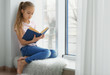 © Africa Studio - Cute little girl with book sitting on windowsill at home