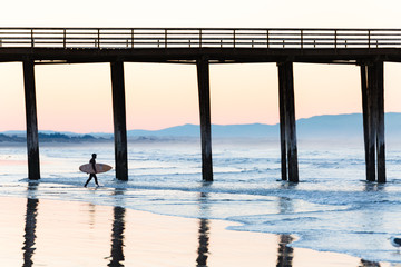 Surfer with surfboard and wetsuit walks towards the water at sunrise in Pismo Beach