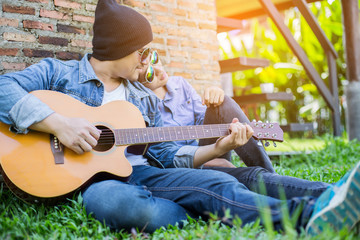 Wall Mural - Hipster man playing guitar for his girlfriend outdoor against br