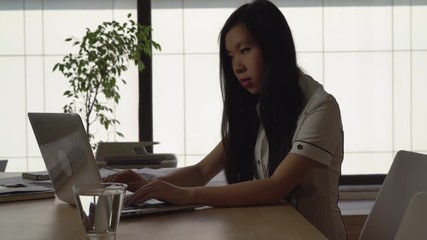 Poster - Young asian woman sitting in the office working with computer. Lady wearing in elegant formal shirt in room with big panoramic windows.