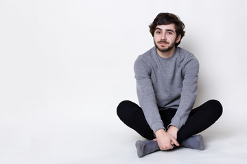 A man with dark beard and moustache having trendy hairstyle wearing casual trousers and grey sweater sits relaxing on the floor with his legs crossed over white background. A stylish hipster.