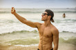 © theartofphoto - Half body shot of a handsome young man using cell phone to take photo, standing on a beach, shirtless wearing boxer shorts and baseball hat, showing muscular fit body