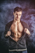 Confident, attractive young man opening vest on muscular torso, ripped abs and pecs. On dark smoky background