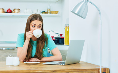 Wall Mural - Young woman working in kitchen with laptop drinking coffee.