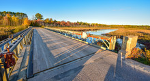 Wide Angle View Of Clear Blue Sky, Late Afternoon Sunshine, Casts Long Shadows On A Light Steel And Timber Bridge, Single Point Perspective Over A Lake In Ontario, Canada.
