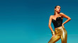 Fashionable girl with a beautiful tan is against the blue clear sky. Girl dressed in a black corset and gold pants. A woman in model pose outdoors. Gold ornaments and bracelets. Promotional poster.