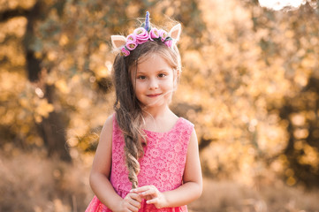 Smiling kid girl 4-5 year old with long hair wearing pink dress and unicorn headband outdoors. Looking at camera. Childhood.