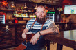 © kanashkin - Bearded barman with tattoos wearing an apron sitting at the bar and holding a glass of beer.