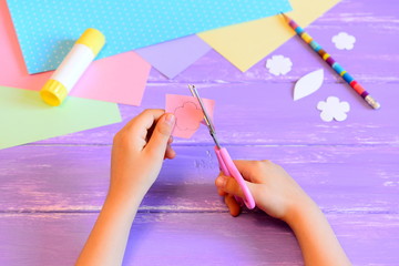 Wall Mural - Small kid makes a greeting card for mom. Kid holds scissors in hand and cuts a flower from paper. Colored paper, paper templates, glue stick, pencil on a table. Mother's day or March 8 crafts concept