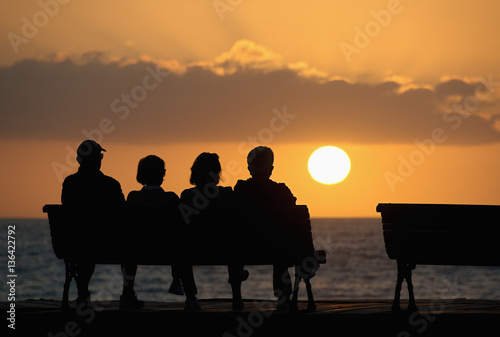 Fotografía Silhouette of a group of seniors who sits on the bench, looking at the sunset
