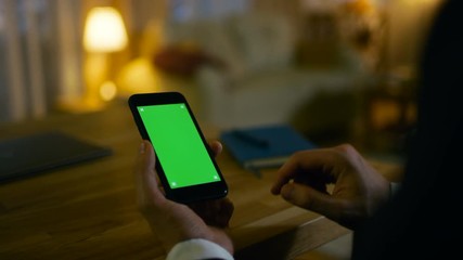 Aufkleber - At Home Man Sits at His Desk and Uses Smartphone with Green Screen on It. He Drinks from a Cup. His Apartment is Done in Yellow colours and is Warm. Shot on RED EPIC-W 8K Helium Cinema Camera.