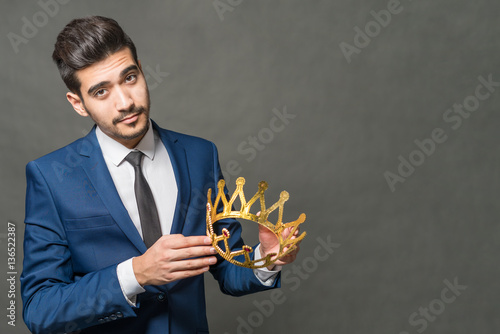 Fotografie, Obraz Young attractive man in a blue suit holding in the hands of the crown on a gray