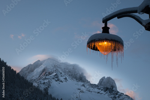 Fotografia, Obraz Snow and icicle covered amber lightpost against backdrop of wind