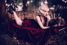 Beautiful And Elegant Blonde Woman With Red Lips And Hair Waves Wearing Wine Red Nightie Posing On The Bed Outdoors Autumn, Retro Vintage Style And Fashion. Retouched Toning Shot