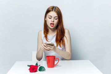 beautiful woman in bright clothes looks at the phone and a cup with a rose on the table