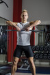 Handsome young man training shoulders lifting barbell, standing in a gym