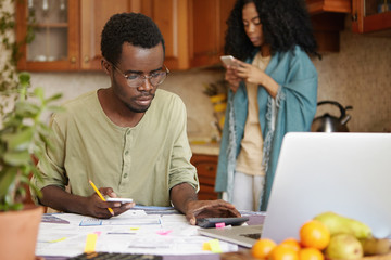 Serious young dark-skinned male in spectacles using cell phone and calculator while calculating family expenses, trying to save some money to buy new car, sitting in front of open laptop computer