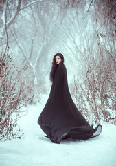 Gothic woman fantasy witch. sexy girl demon walks in forest. wearing a long, black traveling cloak. fallen angel. Dark Queen, with aggressive face make-up. trees covered frost, snow, winter nature