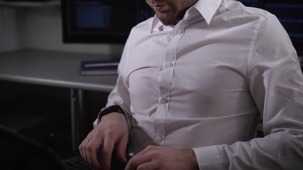 Poster - Close up of a young businessman working on laptop. IT specialist of company is sitting in the office in front of computer monitors, typing on keyboard and looking at the camera.